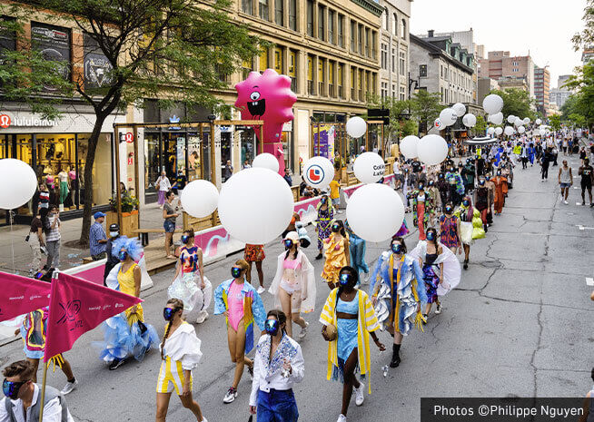 Une parade de rue animée avec des participants en costumes colorés portant de grands ballons blancs, créant une ambiance festive.