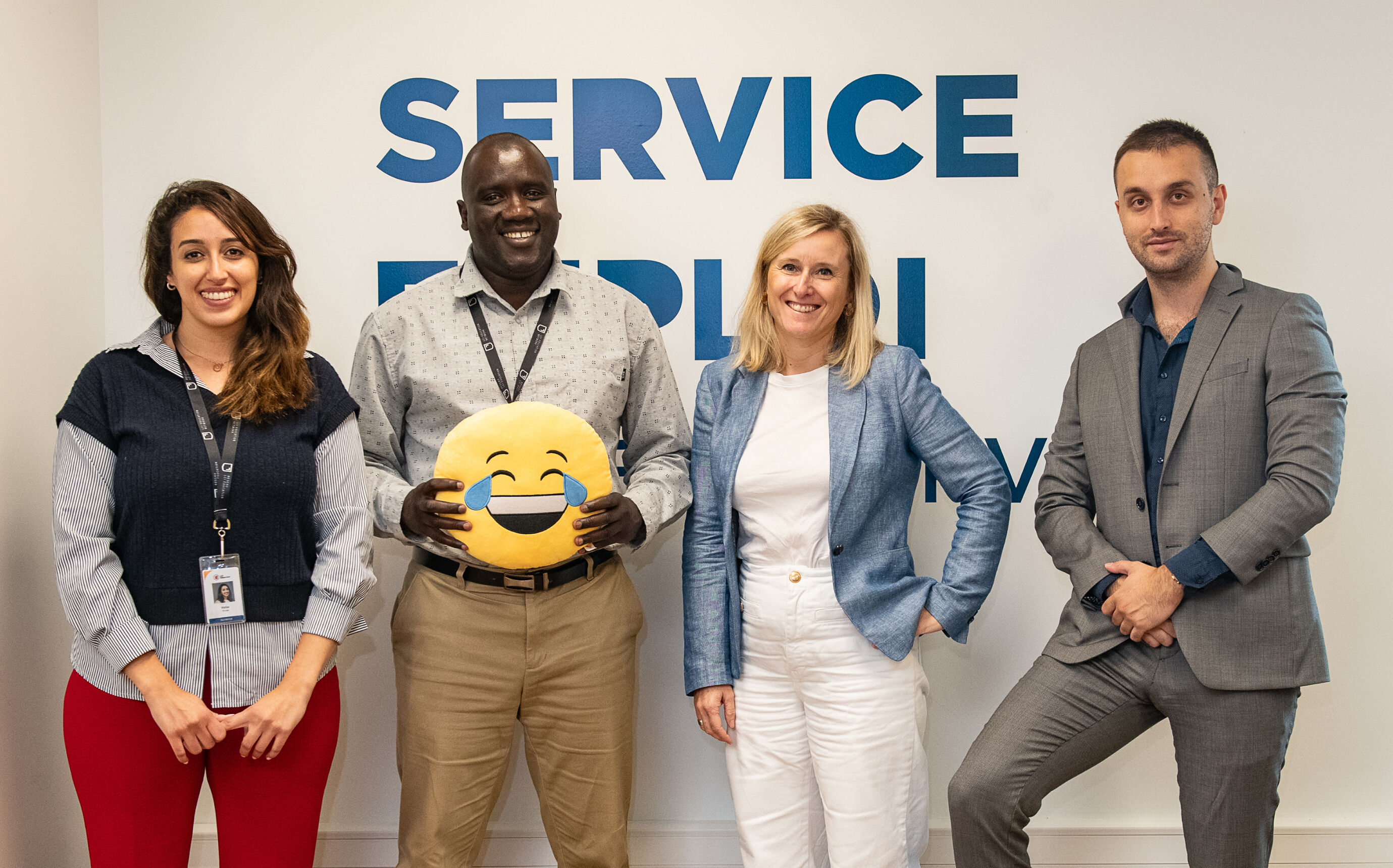 A diverse group of colleagues poses with a laughing emoji cushion, showcasing a positive work environment.