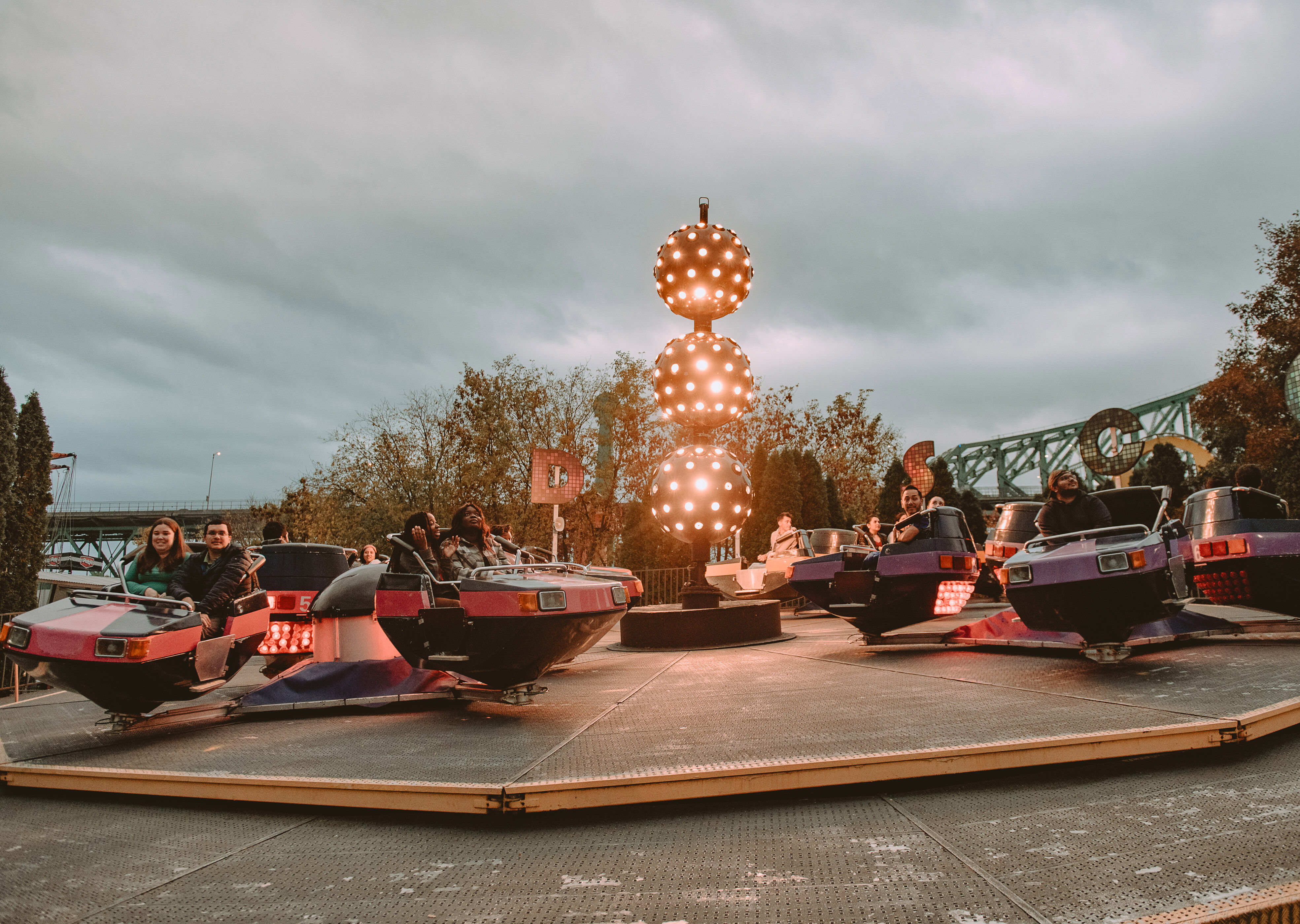Visitors enjoy a bumper car attraction at dusk, with illuminated spherical lights and a backdrop of trees and a roller coaster.
