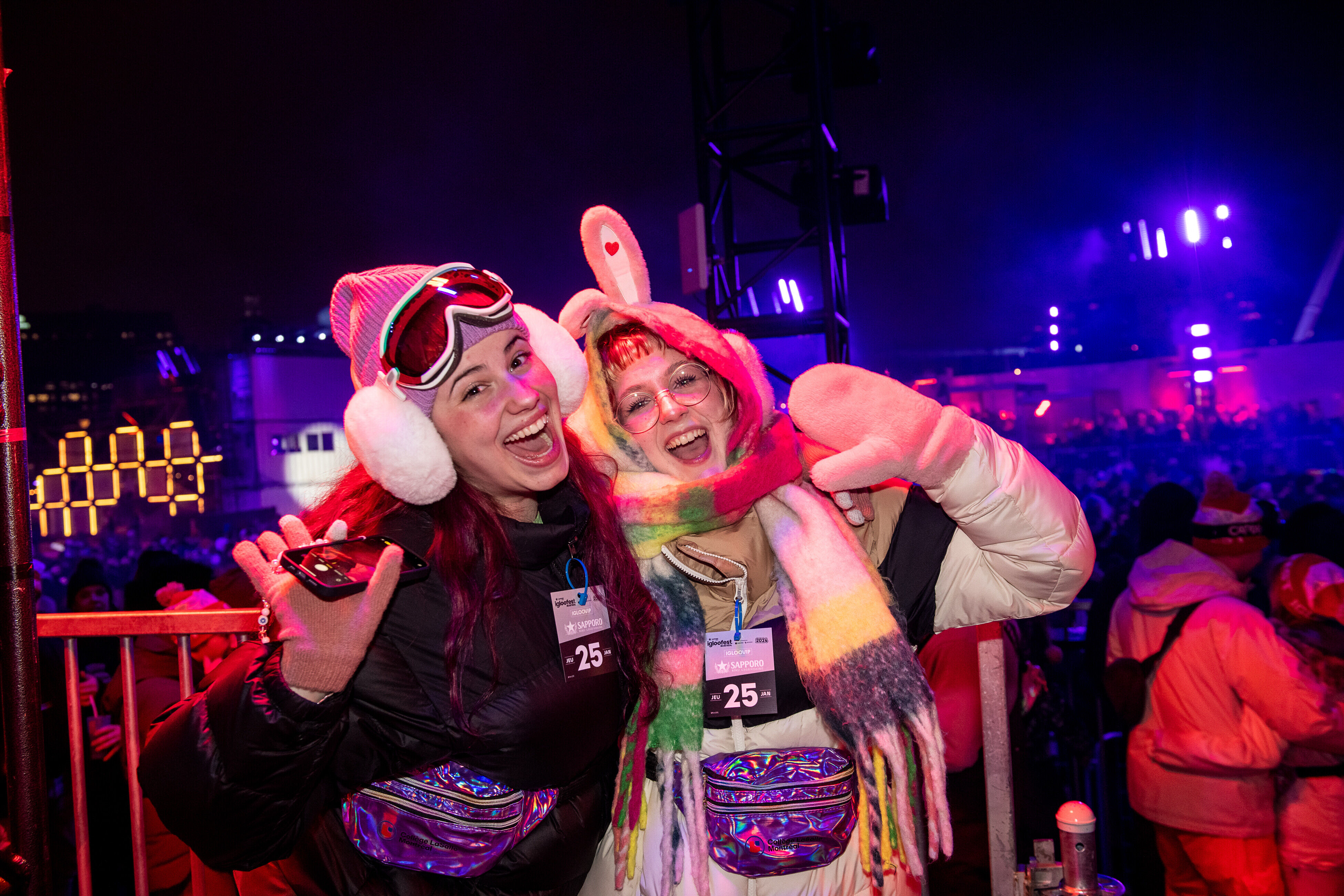 Two women in vibrant winter outfits, with one wearing ski goggles and earmuffs, the other with a colorful scarf, are smiling joyfully at a nighttime outdoor event.