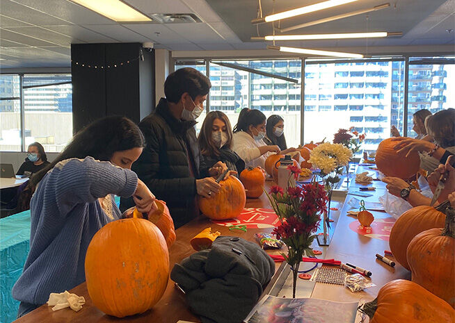 Individuals participating in a pumpkin carving event in a brightly lit indoor setting, with autumnal decorations around.