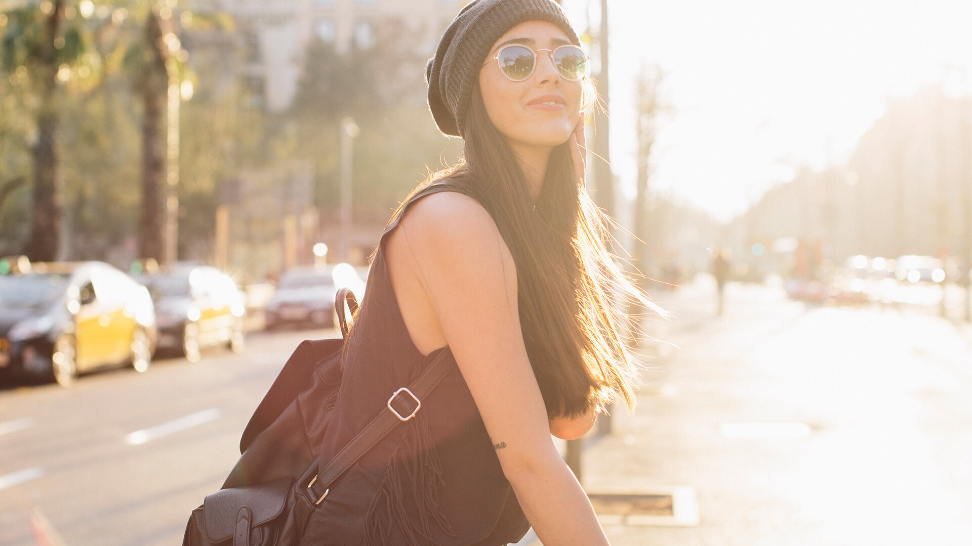 A stylish woman with sunglasses and beanie walks on a sunny city street, radiating a relaxed, modern vibe."