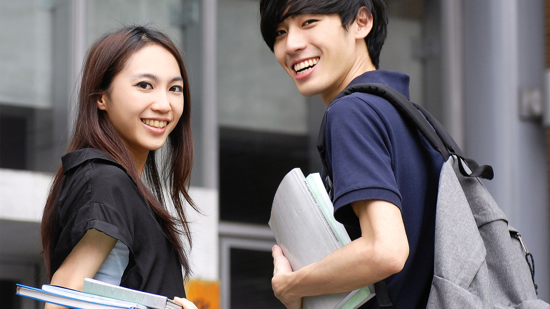 Two happy students with backpacks and textbooks walking outside a campus building.