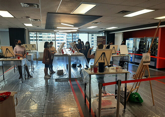 A group of people participating in an art workshop, painting on easels in a room with protective floor coverings.