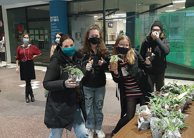 Group of happy individuals with face masks holding potted plants indoors, promoting green living.