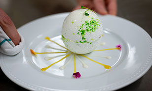 Un dessert présenté de manière complexe qui ressemble à une sphère, orné de garnitures colorées sur une assiette blanche.