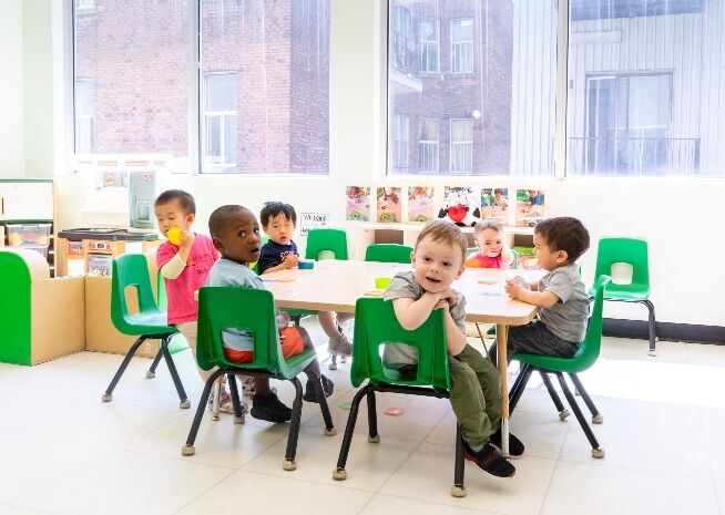 A group of diverse preschoolers sitting at a table, enjoying snack time together in a bright classroom.
