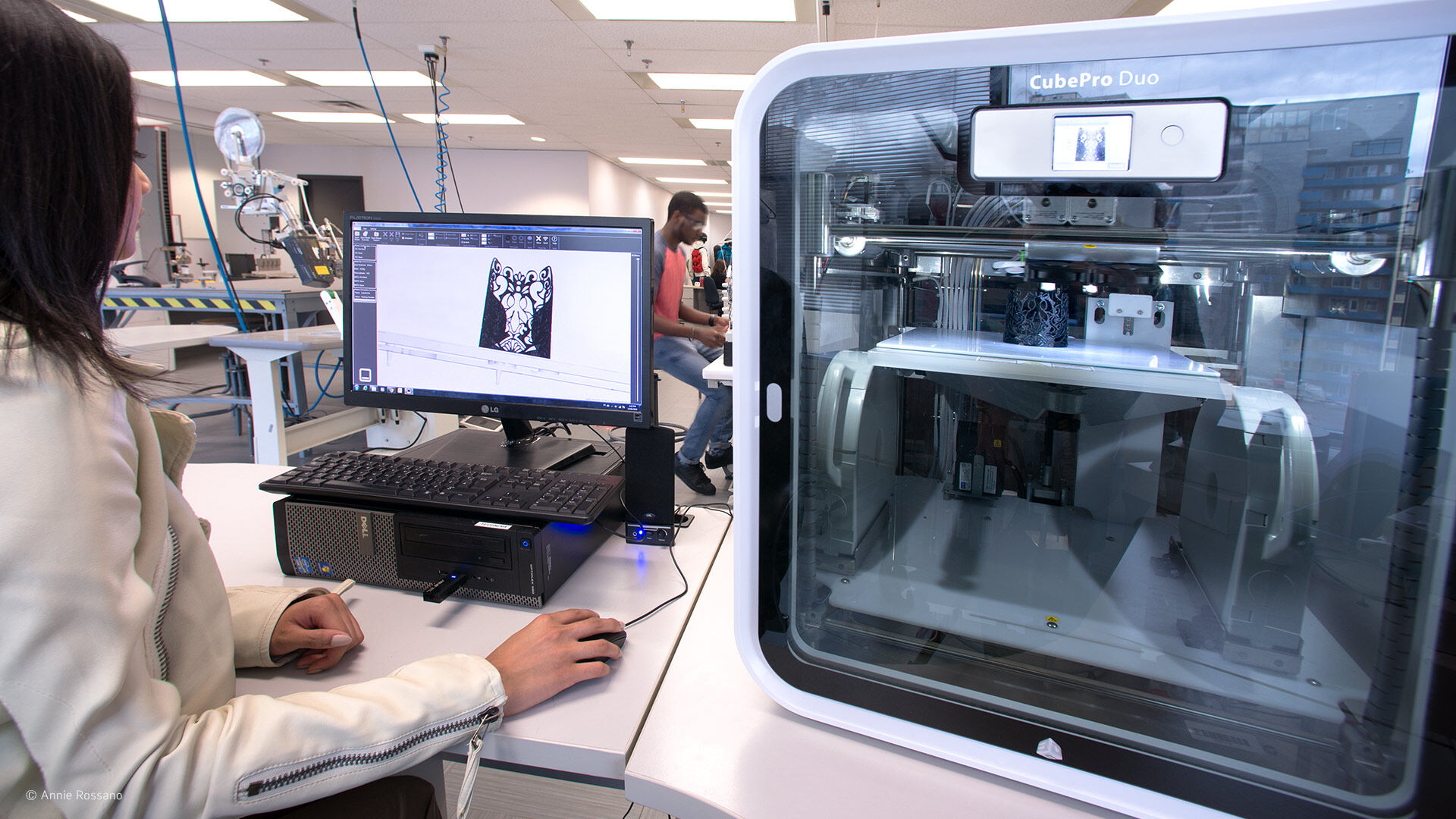A designer finalizes a 3D model on a computer, with a 3D printer in the foreground printing a prototype.