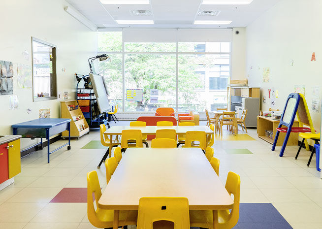 Grande salle de classe de maternelle avec tables et chaises jaunes et divers matériaux éducatifs, éclairée par la lumière naturelle.