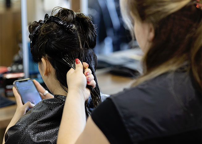 A hairstylist working on a client’s updo, with the client using a smartphone, in a salon environment.