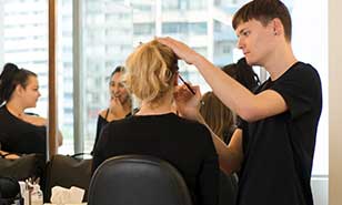 Un jeune coiffeur coiffe méticuleusement les cheveux d'un client dans un salon, avec un miroir reflétant l'intérieur animé.

