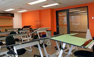 Salle de classe contemporaine avec tables à dessin individuelles et chaises noires, murs orange vifs.