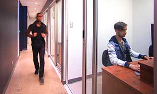 Un homme marche dans un couloir devant une réception où un autre homme est assis derrière un bureau.