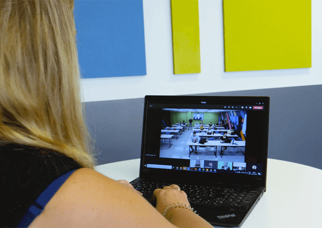 A person observes a classroom through a video conference platform on a laptop, with a colorful backdrop.