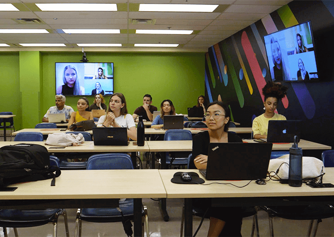 Étudiants en salle de classe avec ordinateurs portables, participant à une session d'apprentissage hybride avec des pairs à distance sur un écran.