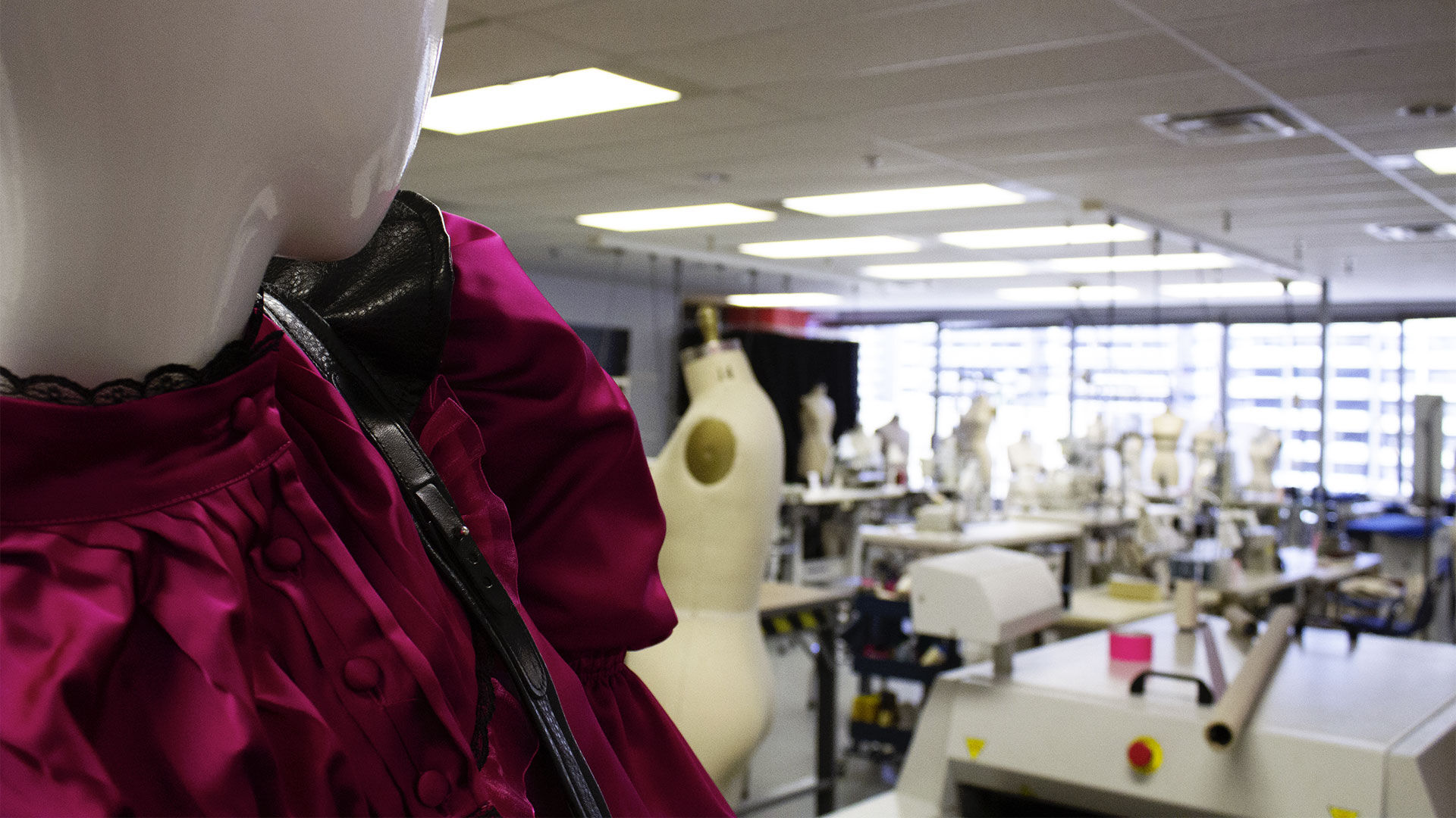 Mannequin in a design studio, wearing a garment in progress, with sewing stations and dress forms in the background.