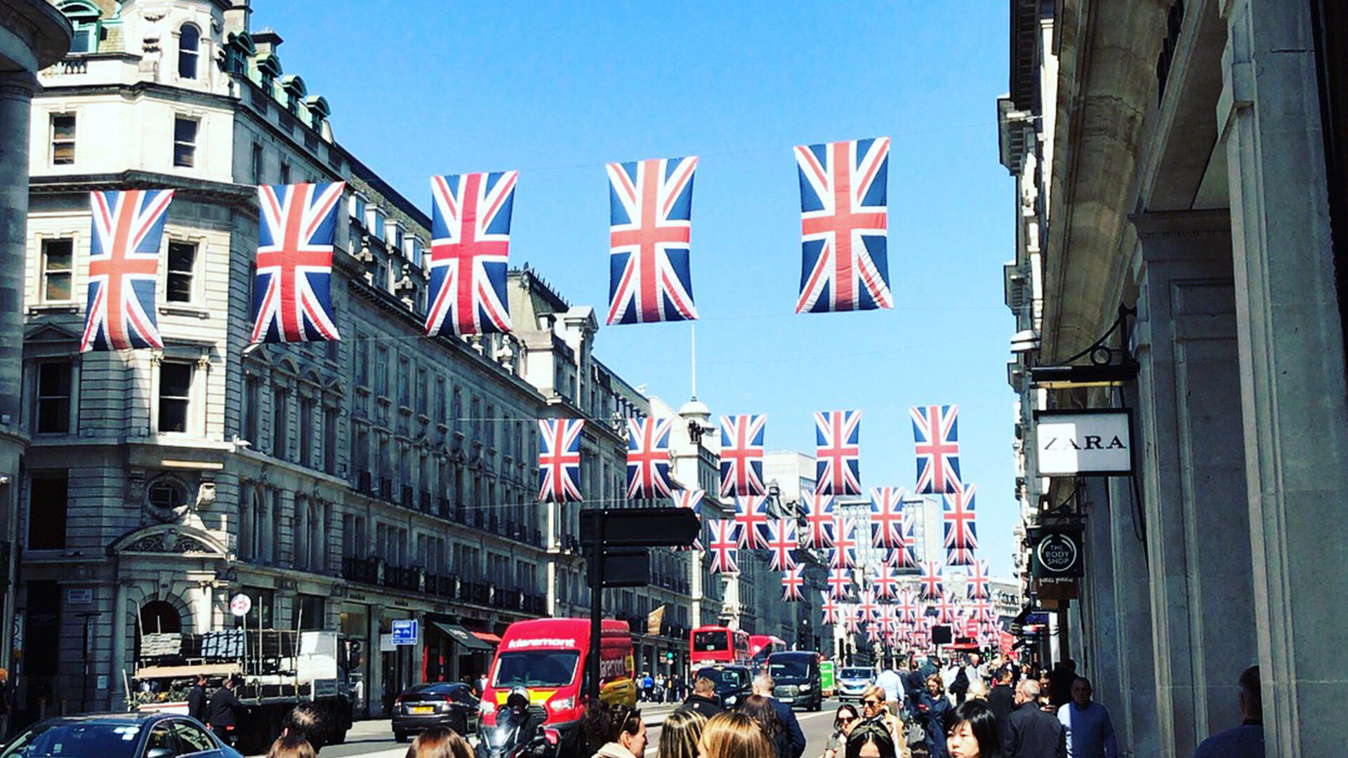 Des drapeaux britanniques décorent une rue animée de Londres, indiquant une célébration ou un événement national, sous un ciel bleu dégagé.
