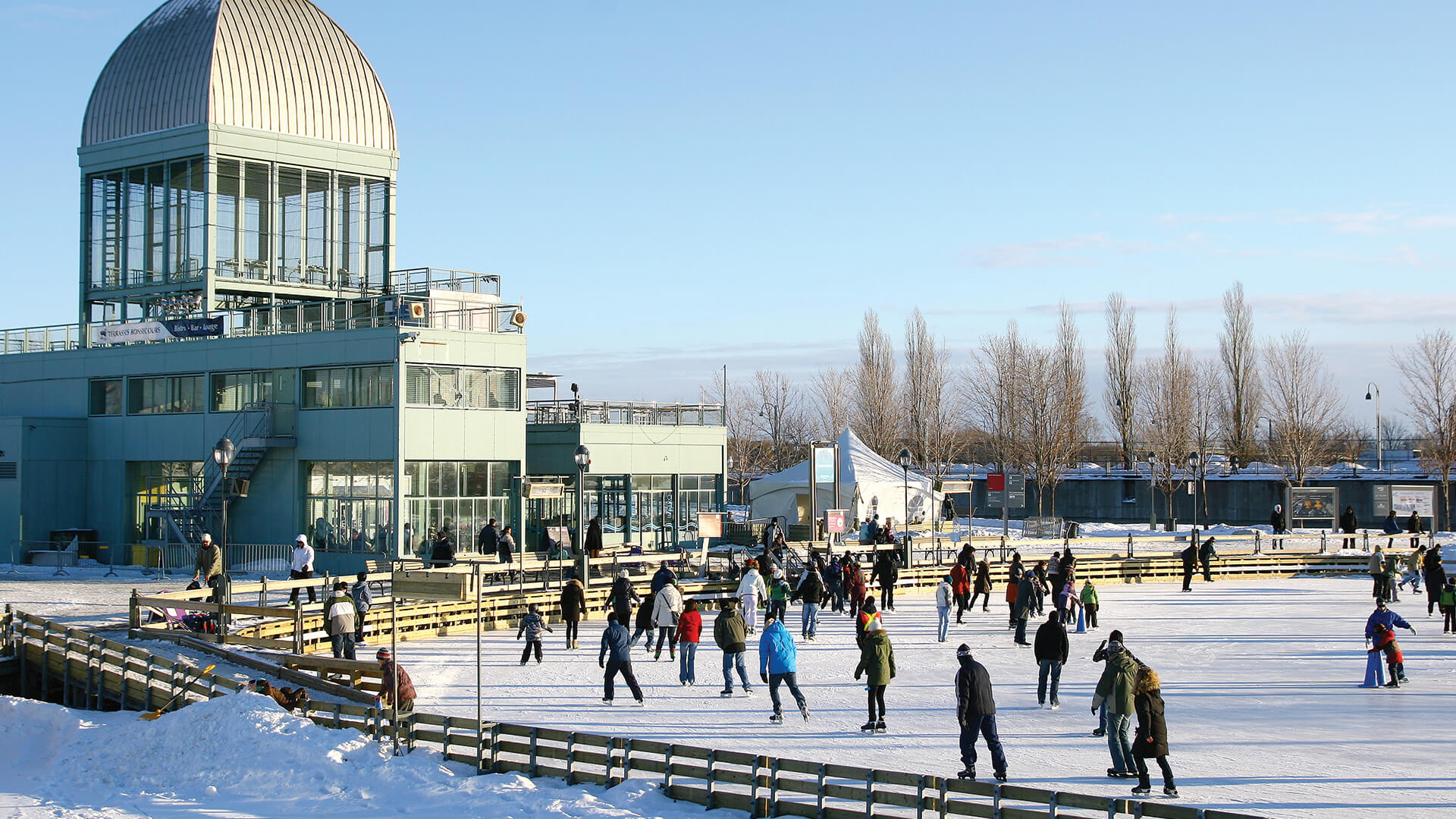People of various ages enjoy ice skating on a large outdoor rink beside a modern building under a clear blue sky.