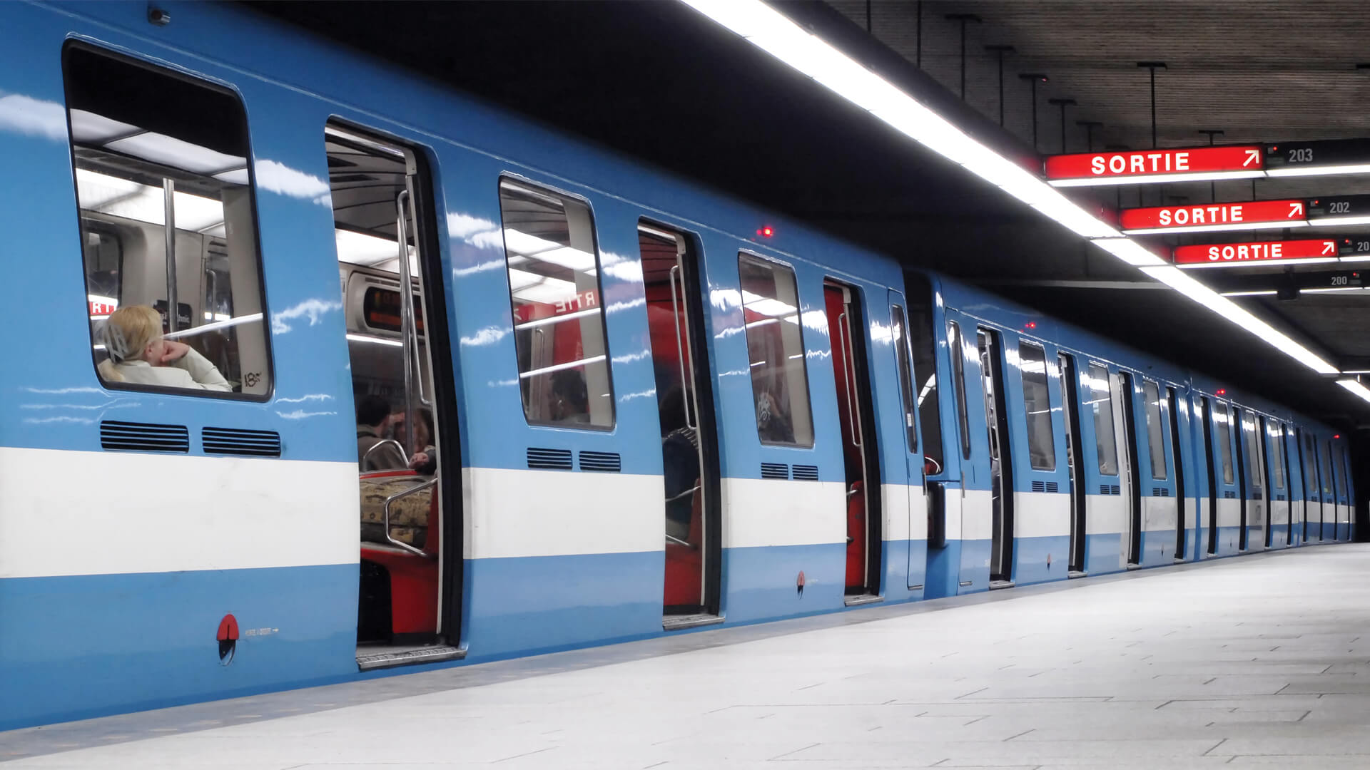 Un train bleu et blanc est à l'arrêt dans une station souterraine éclairée, avec le mot "SORTIE" indiquant les sorties.