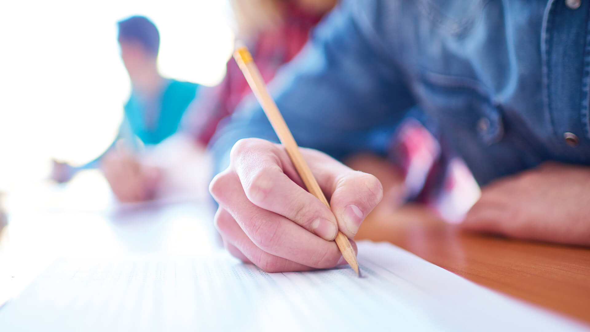 Gros plan sur la main d'un étudiant tenant un crayon au-dessus d'une feuille pendant un examen, avec des camarades en arrière-plan.