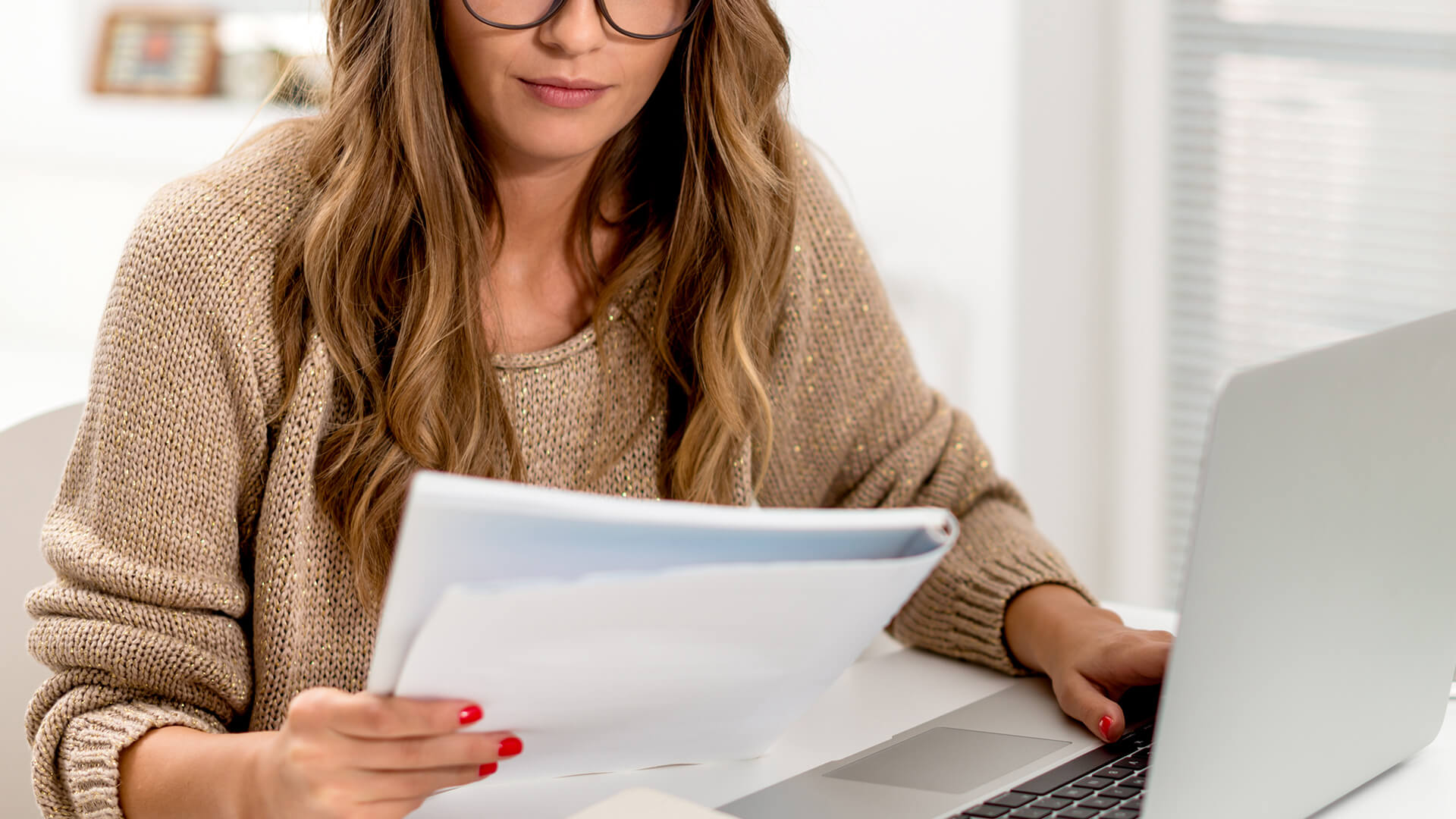 A focused woman with glasses reviews documents beside her laptop in a bright home office.