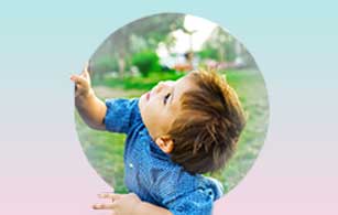 A young child in a blue shirt looks up with curiosity and wonder, arms outstretched, in a park setting.