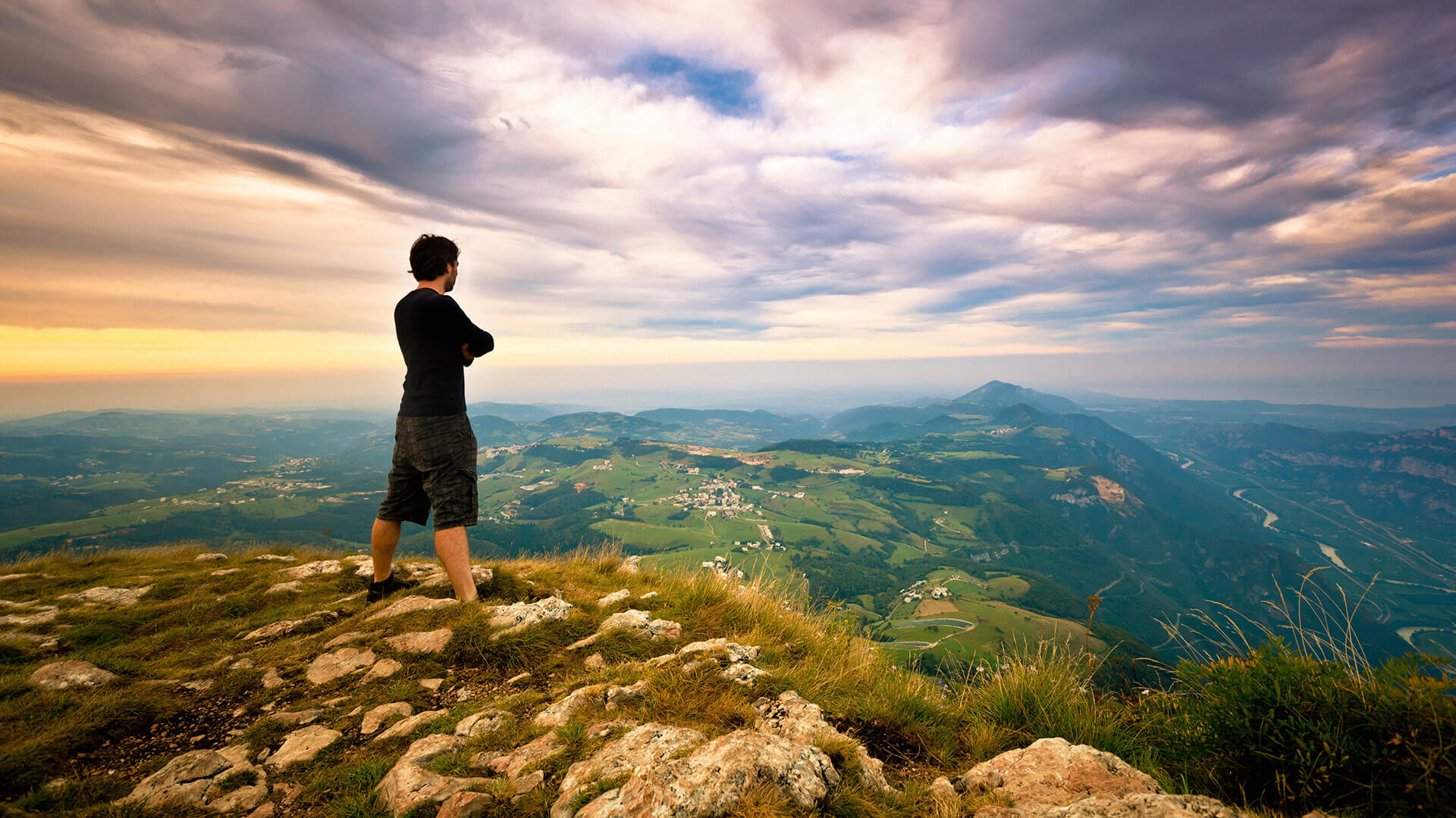 A solitary man stands atop a hill, gazing at a verdant valley under a dramatic sky at dusk, embodying a moment of reflection and vastness.