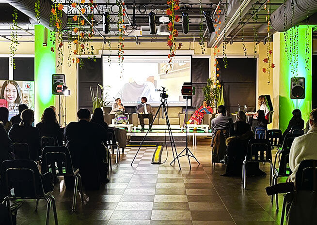 An indoor conference setting with attendees seated facing a stage with speakers, a camera tripod in the aisle, and festive decor.