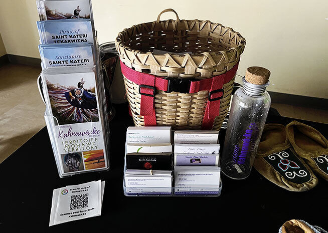 Une table présentant des objets culturels, incluant un panier tissé, des brochures d'information, des cartes nominatives, un QR code, une bouteille d'eau et une pochette en tissu.