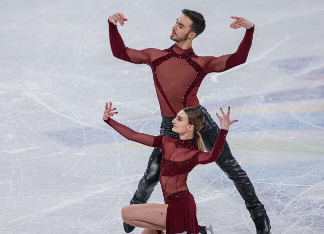 A pair of ice dancers in coordinated burgundy costumes perform a graceful lift.