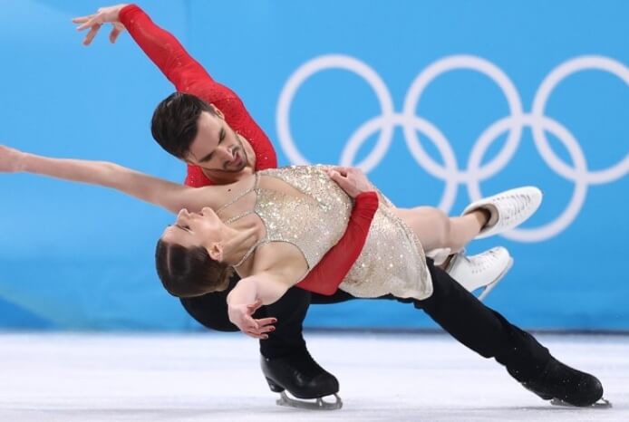 An ice-skating pair in sparkling costumes exhibits a dramatic pose during their routine.