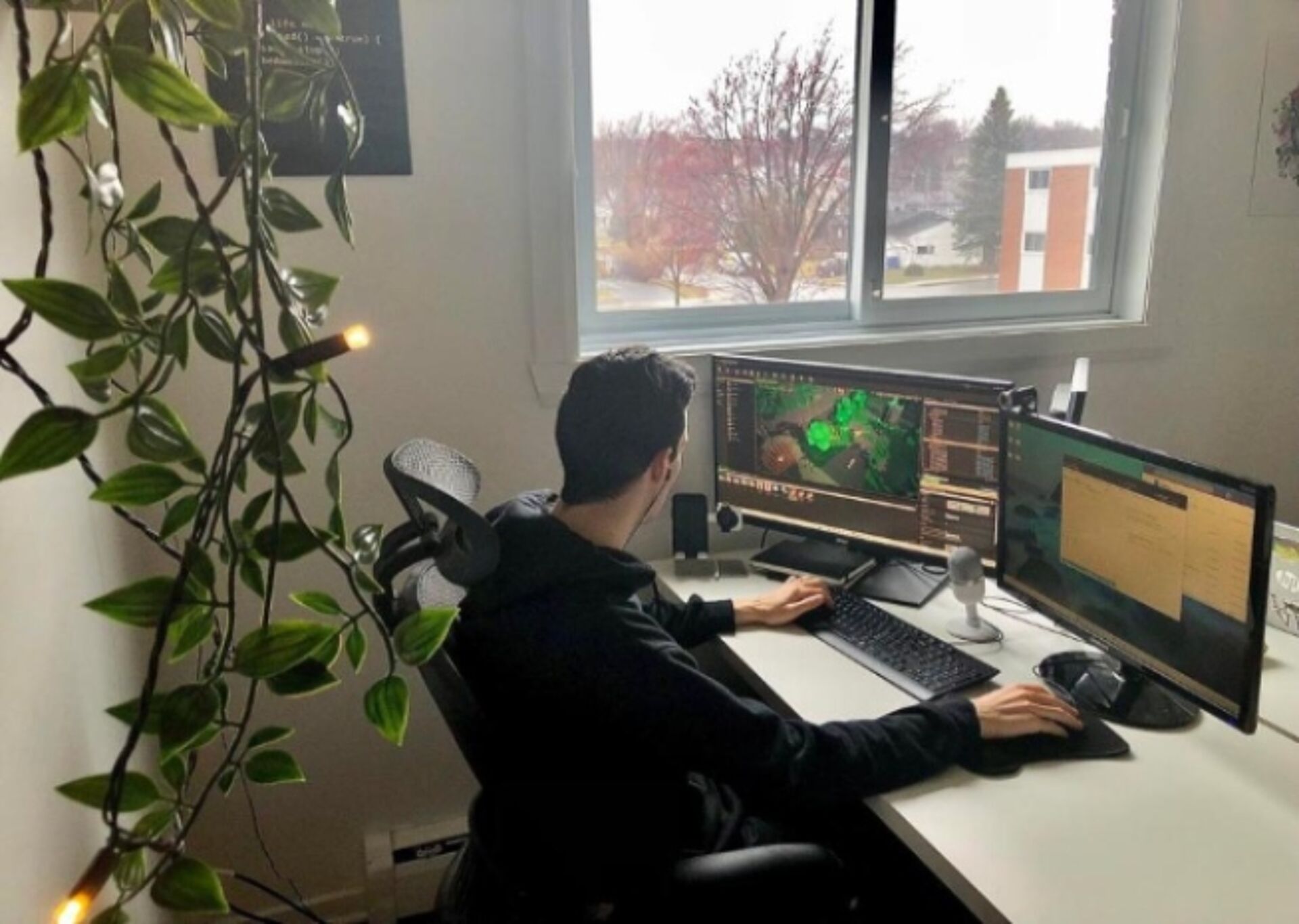 A programmer works intently in a well-lit home office setup with dual monitors and a view outside.