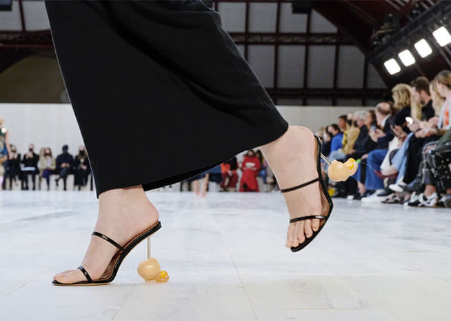 A close-up of a model's foot on the catwalk, wearing a sandal with a quirky heel shaped like a rubber duck.