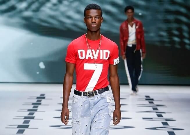 Un mannequin masculin défile sur un podium de mode, portant un t-shirt rouge avec l'inscription "DAVID 7" et un jean délavé clair.