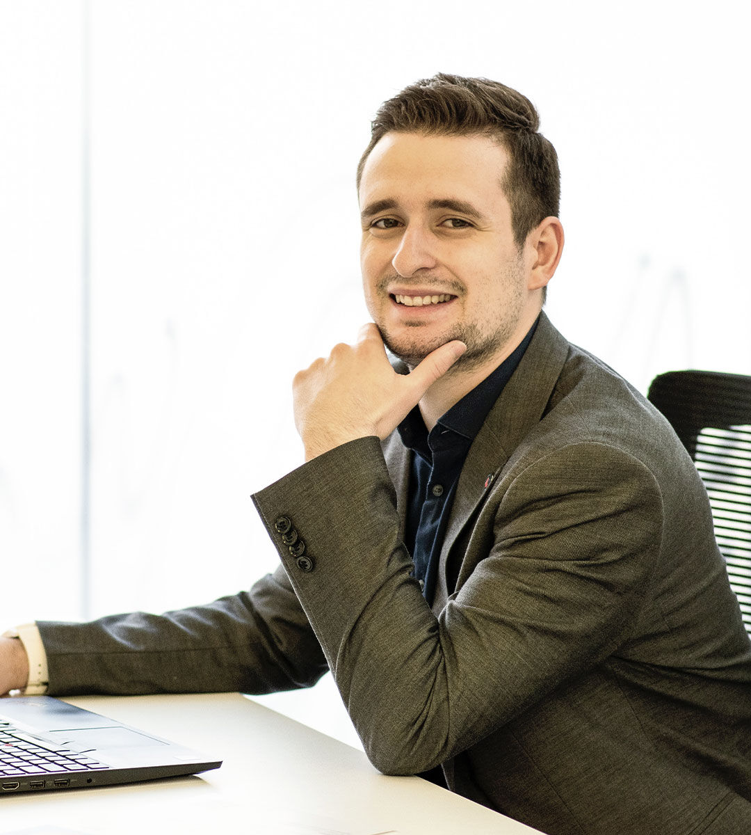 A poised young man with a charming smile, dressed in a smart casual gray blazer, poses at his work desk.