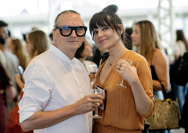 Homme en chemise blanche et grandes lunettes avec un verre de vin, debout à côté d'une femme en blouse rouille tenant également un verre.