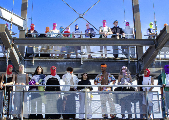 Un groupe hétérogène de spectateurs lors d'un défilé de mode, assis sur un échafaudage métallique, avec des couvre-chefs colorés et des tenues éclectiques.