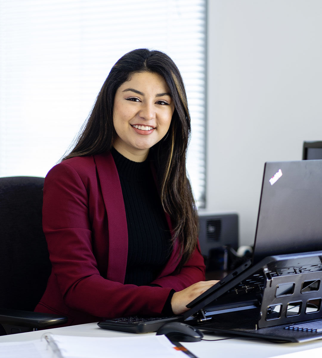 Une femme souriante en blazer rouge assise à un bureau avec un ordinateur portable, incarnant confiance et professionnalisme.