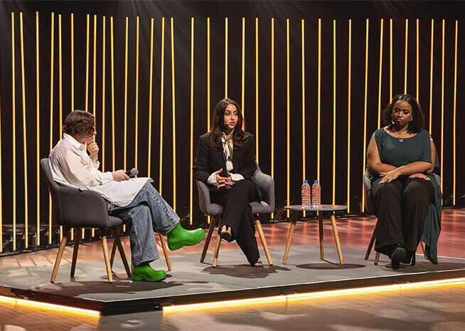 Three people engage in a discussion at a panel event, seated with microphones against a backdrop of vertical light strips.