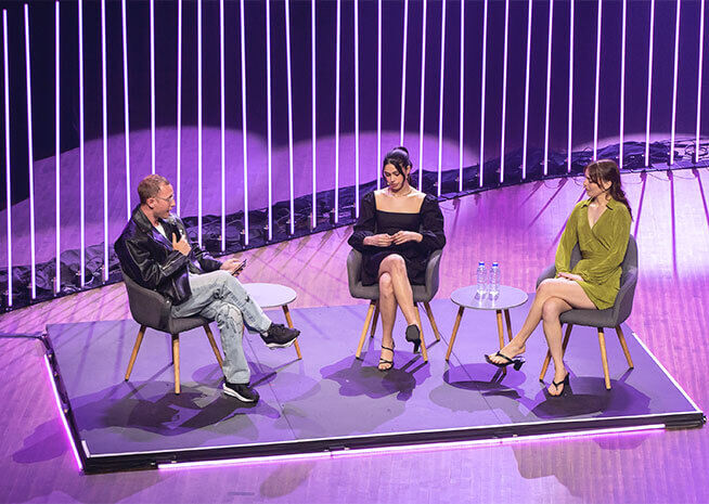 Three people participate in a panel discussion on a stage with vertical lighting accents in the background.