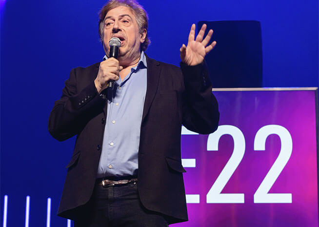 Un homme d'âge moyen avec un microphone s'adresse avec passion à un public lors d'un événement intitulé 'E22'.