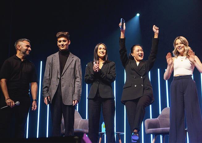 A group of five individuals on stage at an award ceremony, one jubilantly raising her award.