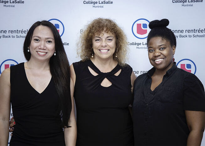 Trois femmes souriantes se tiennent ensemble lors d'un événement éducatif, reflétant la diversité et l'enthousiasme de la communauté académique.