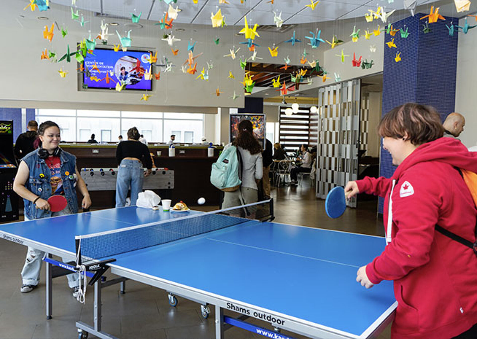 People enjoying a game of ping pong in a relaxed indoor setting with origami decorations overhead.