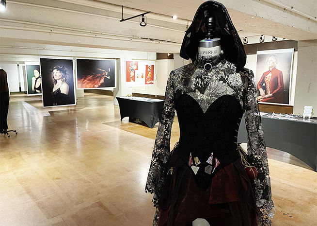 Un mannequin de mode paré d'une robe sombre et complexe se tient devant une galerie de portraits d'art divers.