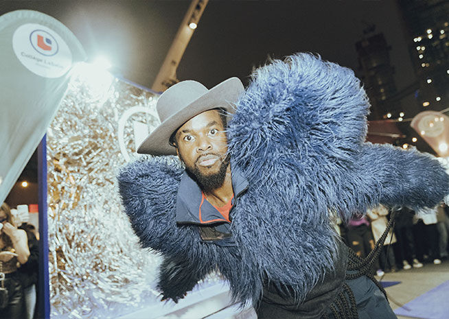 A man strikes a pose in a flamboyant fur coat and hat, exuding confidence amidst a glittering party backdrop.