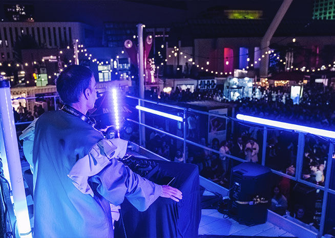 DJ orchestrates the crowd from above, with a sea of partygoers dancing under the neon lights.