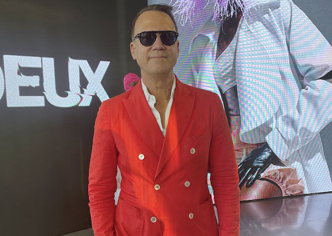 A fashion designer in a bold red blazer stands confidently, with a dynamic digital display in the background.