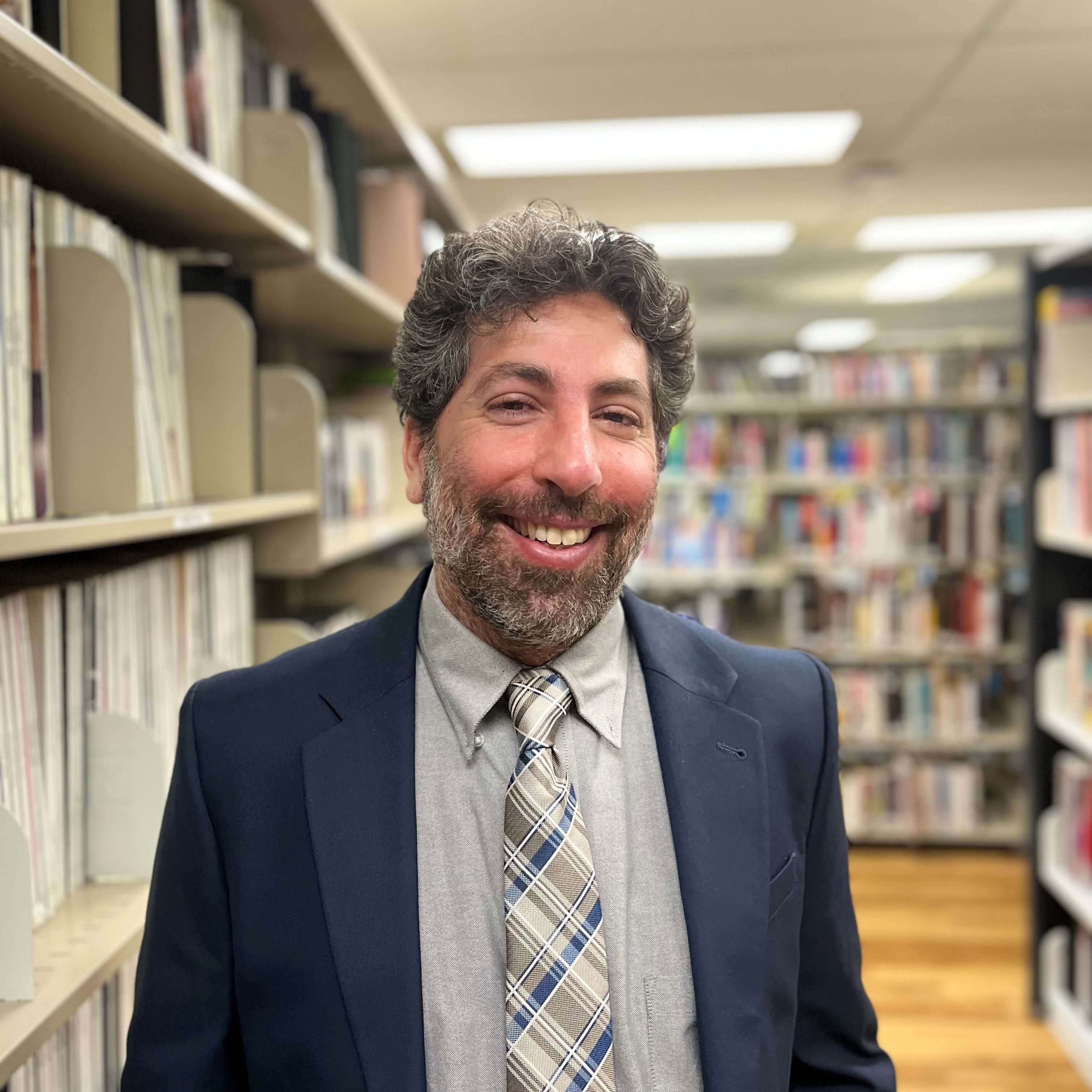 A jovial man with curly gray hair, sporting a blue suit, tie, and a gray shirt, grinning in a library setting with bookshelves in the background.