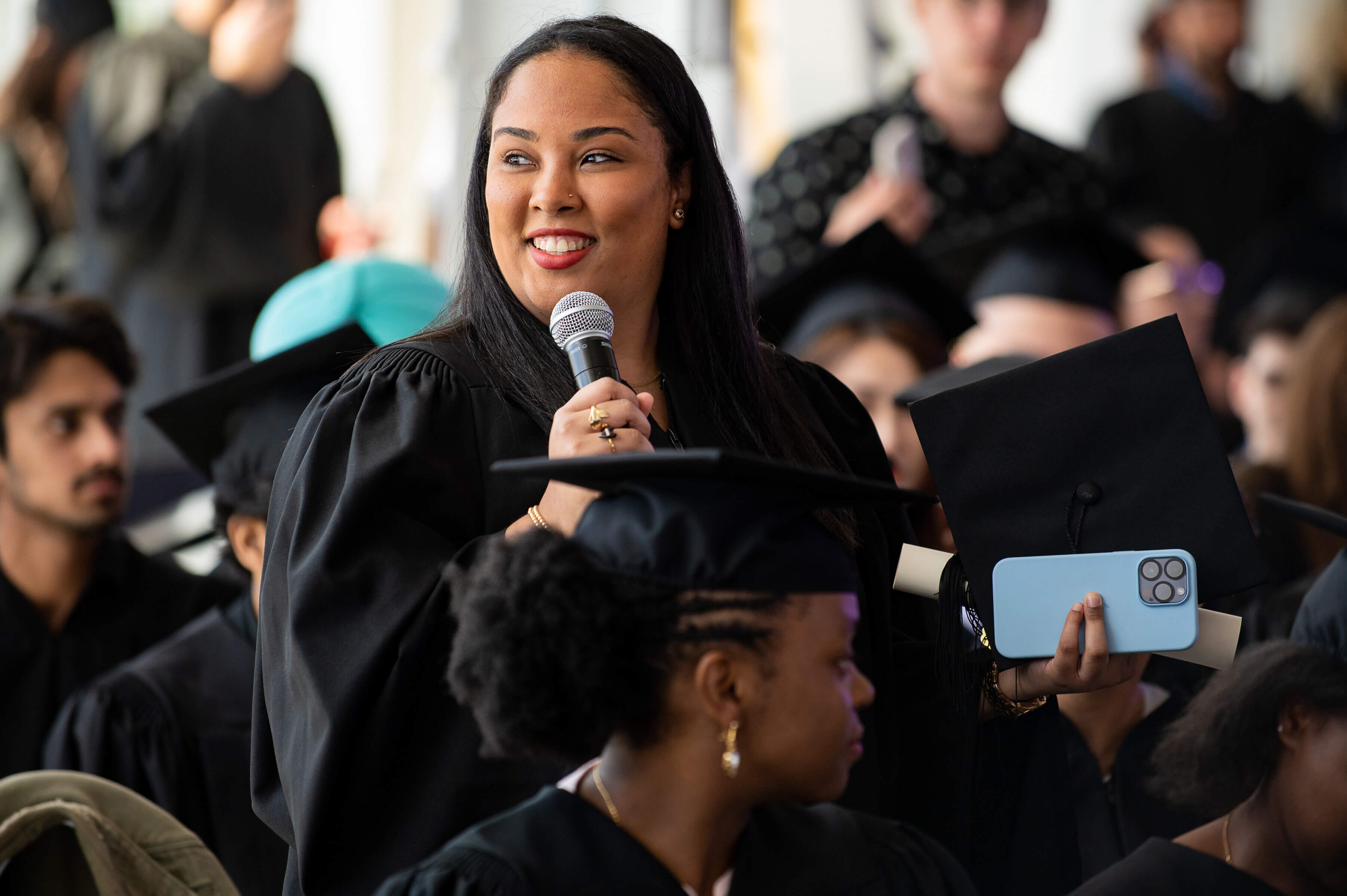 Une femme posée prononce un discours lors de sa remise de diplôme, ses pairs en toges écoutant attentivement.